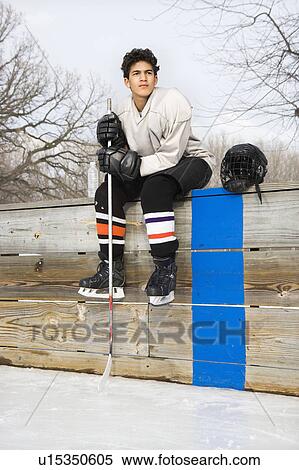 Stock Photography - Boy in ice hockey uniform holding hockey stick sitting on sidelines.. Fotosearch