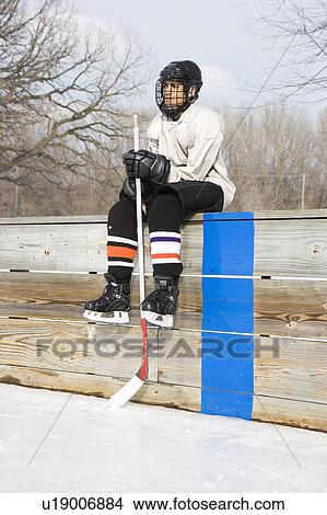 Picture - Hockey player holding hockey stick sitting on sidelines. Fotosearch