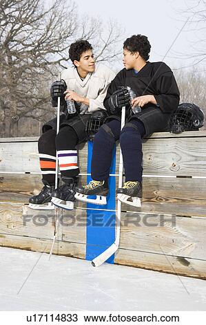 Two boys in hockey uniforms sitting on ice rink wall View Large Photo Image Stock Image - Two boys in hockey uniforms sitting on ice rink wall. Fotosearch