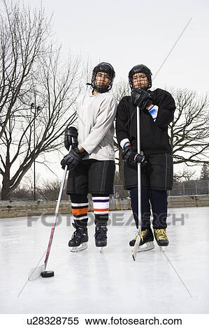 Stock Photography - Two boys in ice hockey uniforms holding hockey sticks standing on ice rink in ice skates.. Fotosearch