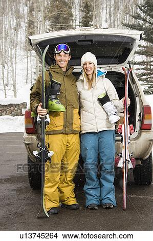 Picture - Young couple wearing winter clothes standing by automobile with ski equipment smiling.. Fotosearch