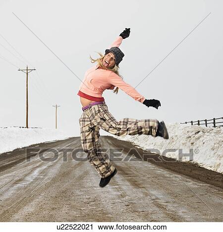 Stock Image - Young woman in winter clothes standing on muddy dirt road jumping and smiling.. Fotosearch