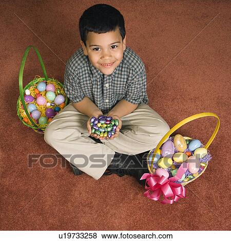 Stock Photo - Hispanic boy sitting on floor with two Easter baskets holding chocolate candy eggs in his hands looking up at viewer smiling.. Fotosearch