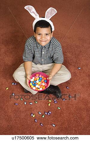 Stock Photography - Hispanic boy wearing bunny ears sitting on floor holding a bowl of chocolate candy eggs with more candy scattered around him looking up at viewer smiling.. Fotosearch