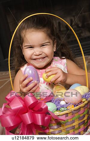 Stock Photo - Hispanic girl holding egg from Easter basket looking at viewer smiling.. Fotosearch