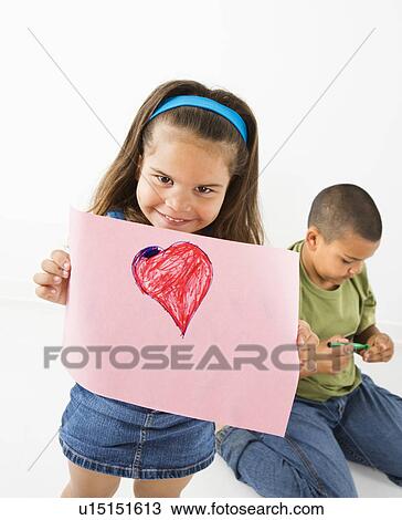 Stock Image - Young girl showing off drawing of heart while boy sits behind her. Fotosearch