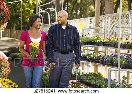 Stock Image - Happy smiling couple picking out flowers at outdoor plant market walking and holding hands.. Fotosearch