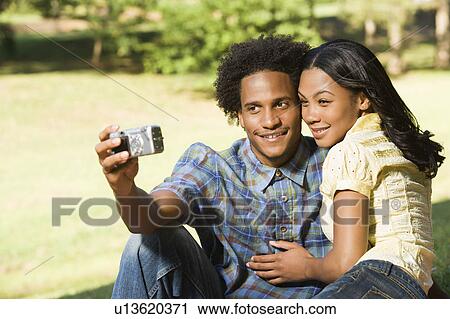 Stock Image - Happy smiling couple taking pictures together in park with digital camera.. Fotosearch