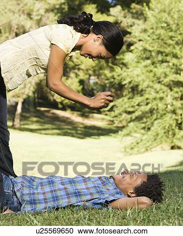 Stock Image - Woman standing over smiling man laying in grass taking his photograph.. Fotosearch