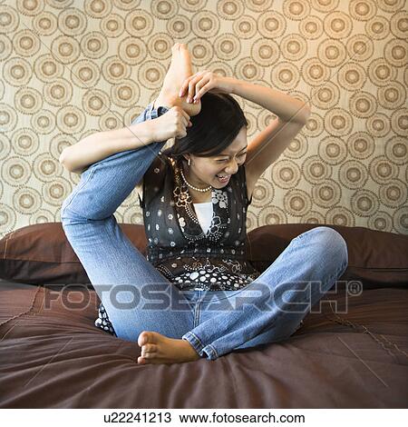 Stock Image - Woman sitting on bed trying to place foot behind her head. Fotosearch