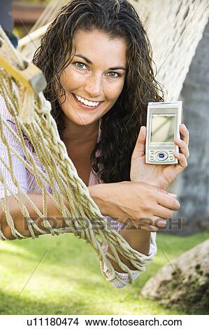 Smiling pretty young adult brunette female laying in hammock holding up PDA towards viewer View Large Photo Image Picture - Smiling pretty young adult brunette female laying in hammock holding up PDA towards viewer. Fotosearch