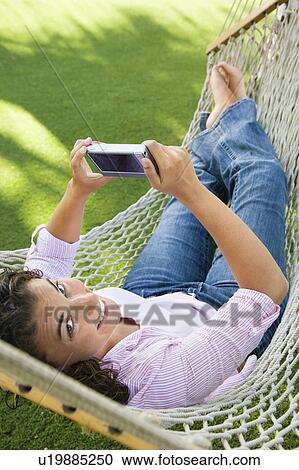 Stock Image - Smiling pretty young adult Caucasian brunette female lying in hammock looking up at veiwer and using PDA.. Fotosearch