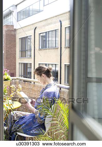 Stock Image - Woman sitting on balcony reading book. Fotosearch