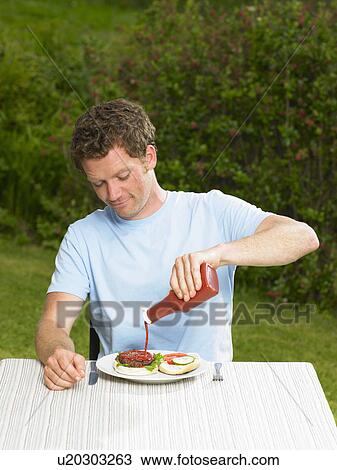 Stock Image - Young man pouring ketchup on burger. Fotosearch