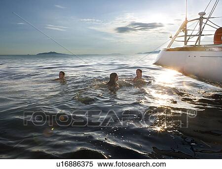 Stock Photography - Three people swimming next to sailboat. Fotosearch