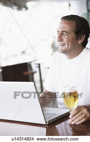 Picture - Mid adult man sitting in front of a laptop and holding a glass of beer. Fotosearch