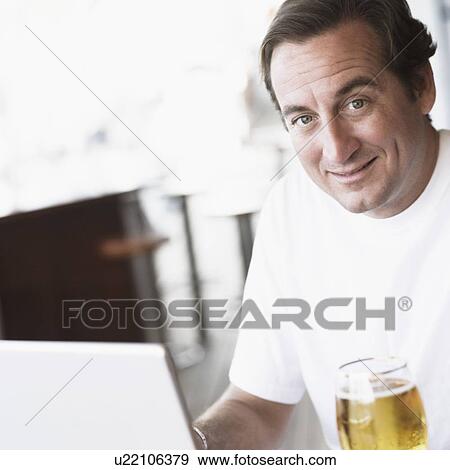 Portrait of a mid adult man sitting in front of a laptop and holding a glass of beer View Large Photo Image Stock Photo - Portrait of a mid adult man sitting in front of a laptop and holding a glass of beer. Fotosearch