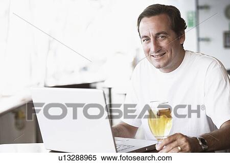 Portrait of a mid adult man sitting in front of a laptop and holding a glass of beer View Large Photo Image Stock Photography - Portrait of a mid adult man sitting in front of a laptop and holding a glass of beer. Fotosearch