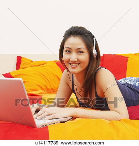 Stock Photo - Portrait of a young woman lying on the bed and using a laptop. Fotosearch