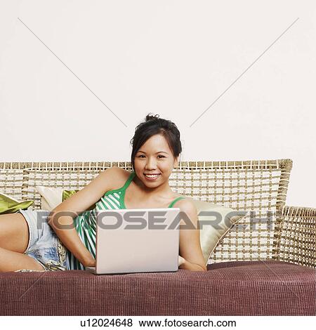 Stock Photo - Portrait of a young woman using a laptop on a couch and smiling. Fotosearch