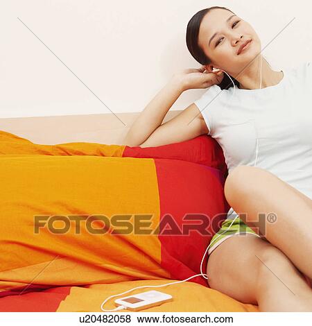 Stock Photo - Portrait of a young woman reclining on the bed and listening to music on headphones. Fotosearch