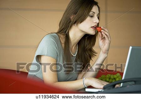 Businesswoman eating a strawberry at laptop View Large Photo Image Picture - Businesswoman eating a strawberry at laptop. Fotosearch