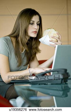 Stock Photograph - Businesswoman eating pita sandwich at laptop. Fotosearch