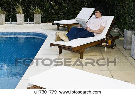 Stock Photo - Young man on chaise lounge by pool reading. Fotosearch