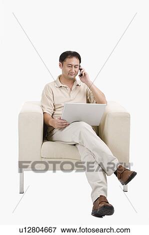 Stock Photo - Businessman sitting on sofa, using laptop and on the phone, Business People. Fotosearch
