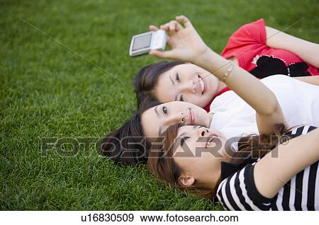 Stock Photo - Three young women lying on lawn and taking pictures. Fotosearch