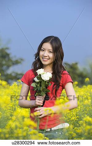 Picture - Young woman holding a bunch of flower and standing in the flower field. Fotosearch