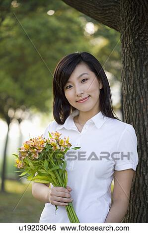 Stock Photograph - Young woman holding a bunch of flowers and leaning on a tree. Fotosearch