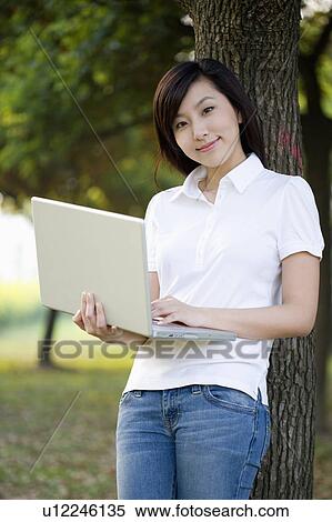 Stock Photography - Young woman using laptop and leaning on a tree, Studying, Education. Fotosearch
