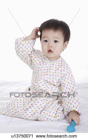 Stock Image - Baby girl sitting with hand in hair and holding a toy, Child. Fotosearch
