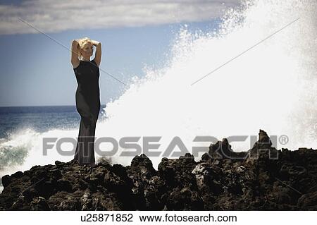 Stock Image - Woman in evening dress standing on the rocks as the surf breaks. Fotosearch
