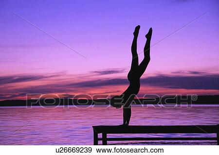 Stock Photo - A person in a hand stand on a dock against a sunset. Fotosearch