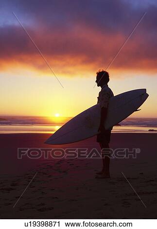 Stock Image - Silhouetted surfer standing on beach at sunset.. Fotosearch