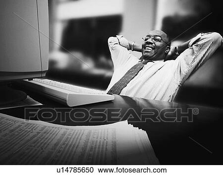Stock Image - Man leaning back in chair in front of desk with computer. Fotosearch