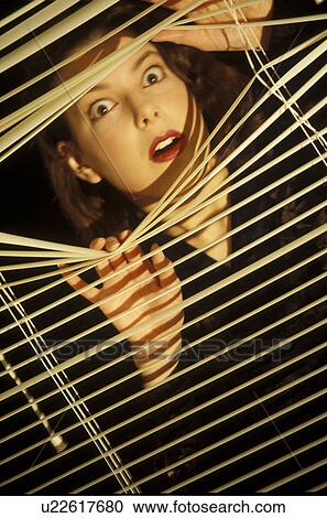 Stock Image - Young woman looking through blinds with surprise. Fotosearch