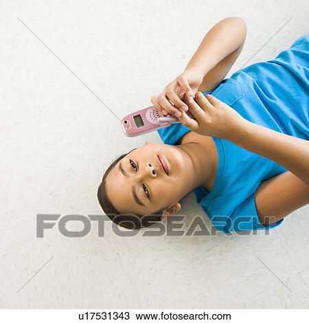 Stock Image - Asian preteen girl lying on back on floor text messaging on cell phone.. Fotosearch