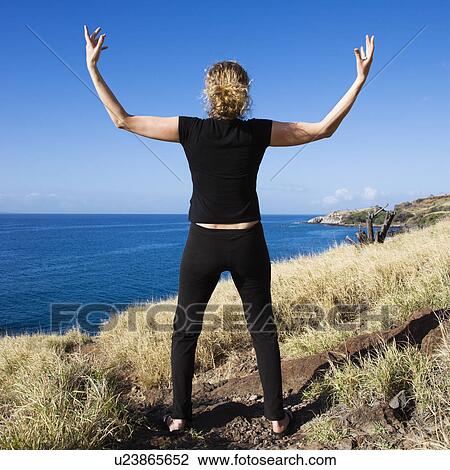 Stock Image - Back view of Caucasian mid-adult woman standing on Maui, Hawaii coast with arms raised above head.. Fotosearch