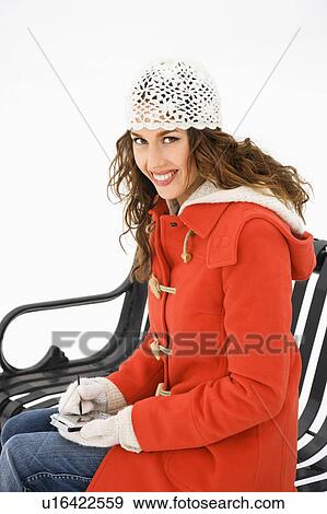 Stock Photo - Caucasian young adult female in winter clothing sitting on park bench using PDA and smiling at viewer.. Fotosearch