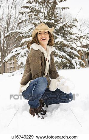 Stock Photo - Caucasian young adult female smiling at viewer while kneeling in snow with snowball and wearing straw cowboy hat.. Fotosearch