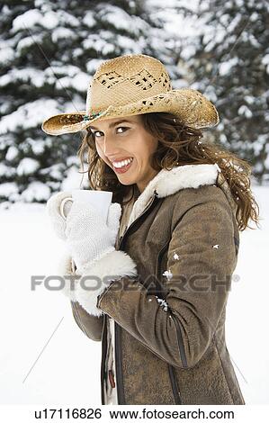 Stock Photograph - Caucasian young adult female wearing straw cowboy hat outdoors in snow holding coffee cup and smililng at viewer.. Fotosearch