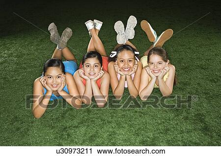 Stock Image - Four multiethnic girls lying on ground in indoor gym with heads on hands smiling.. Fotosearch