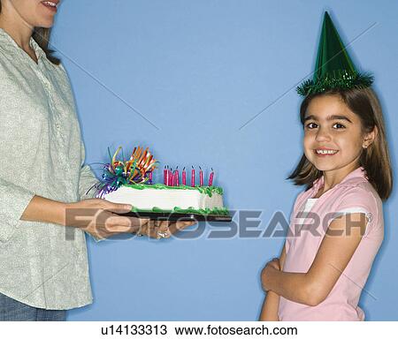 Stock Image - Girl wearing party hat with mother holding birthday cake.. Fotosearch
