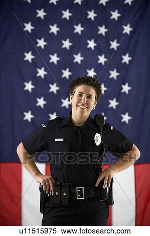 Stock Photography - Portrait of mid adult Caucasian policewoman standing with hands on holster and American flag as backdrop smiling at viewer.. Fotosearch