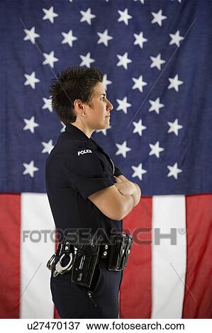 Stock Photo - Side view of mid adult Caucasian policewoman standing with arms crossed and American flag as backdrop.. Fotosearch
