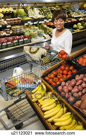 Stock Image - Smiling middle aged African American woman selecting tomatoes from grocery store produce.. Fotosearch