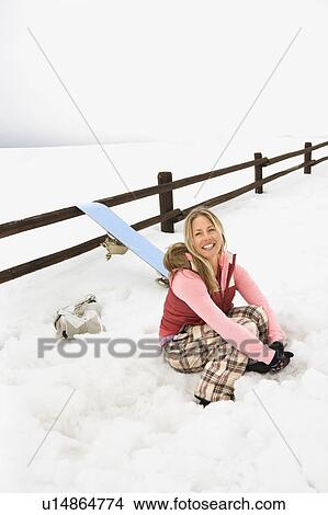 Young woman in winter clothes by fence in snowy field kneeling by snowboard and smiling. View Large Photo Image Picture - Young woman in winter clothes by fence in snowy field kneeling by snowboard and smiling.. Fotosearch
